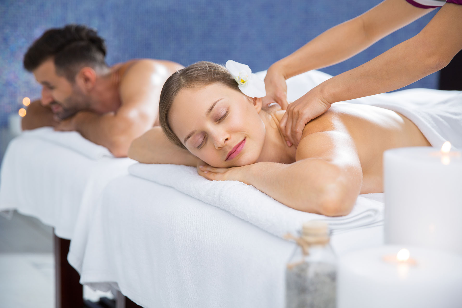 Couple getting massage together in spa center. Young Caucasian woman with flower in hair getting back massage. Man lying on massage table and relaxing after treatment in background